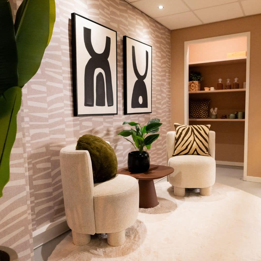 Elle Professionals 2 Overlegruimte in Japandi en bohemian stijl met beige, grijze, bruine en groene accenten. Aangekleed met prachtige fauteuils, een hoogpolig vloerkleed, sierkussens, kunst, kamerplanten en een kast vol gezellige kopjes en potjes.