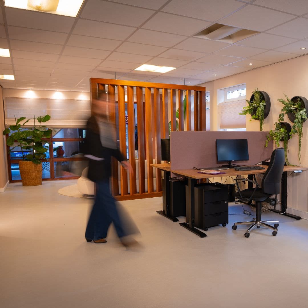 Elle Professionals 3 Kantoorruimte met meerdere werkplekken, gietvloer, scheidingswand, computers, kamerplanten en een persoon in beweging voor een levendige sfeer.